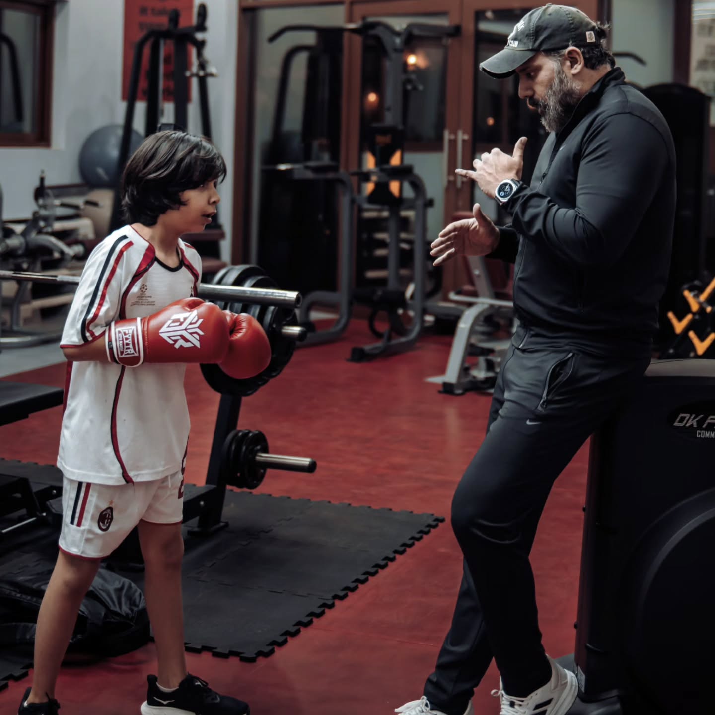 A boy in red boxing gloves listening as an adult in a cap shows him a hand position in a gym