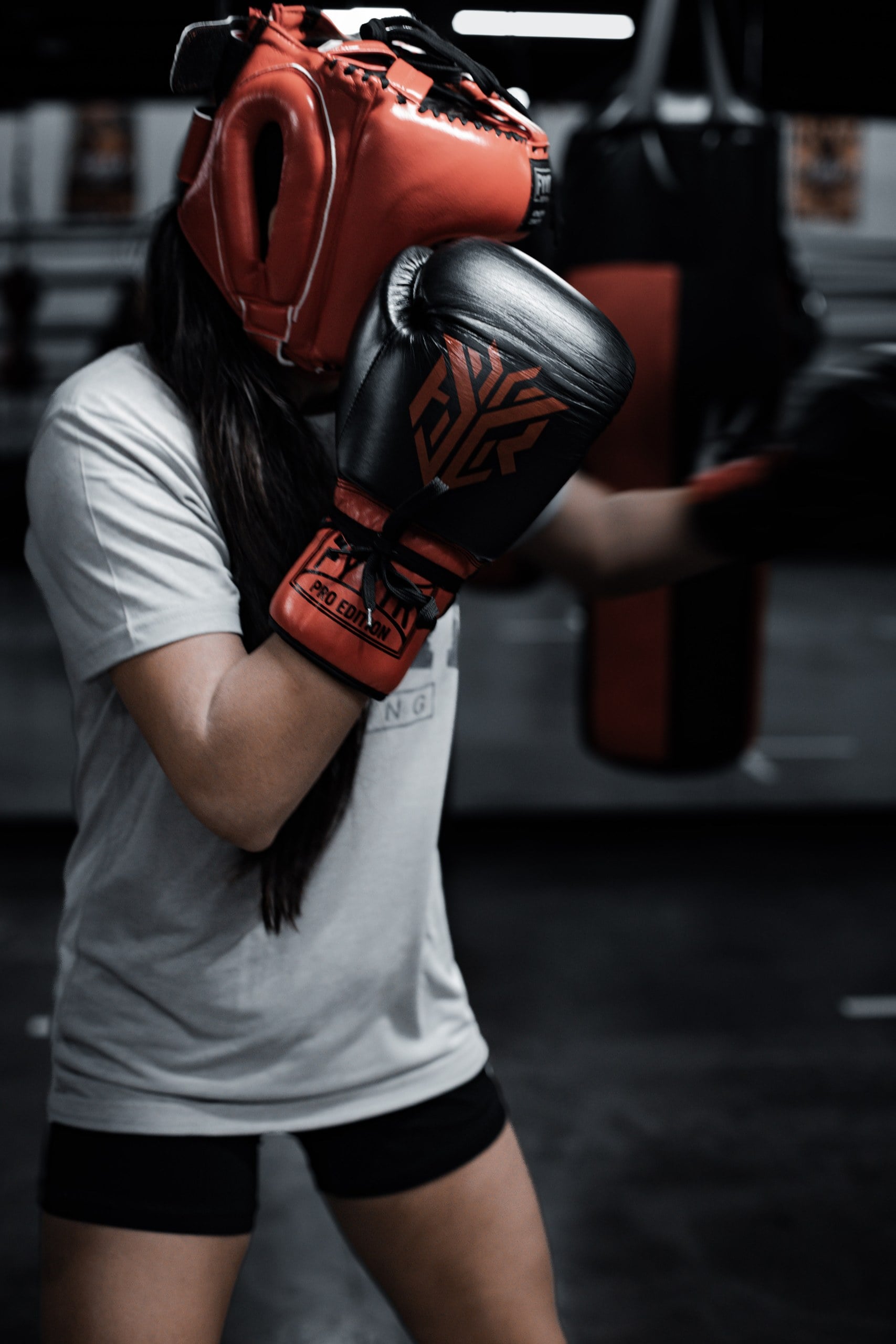Boxer in a red headguard with her guard up in a gym with heavy bags behind her